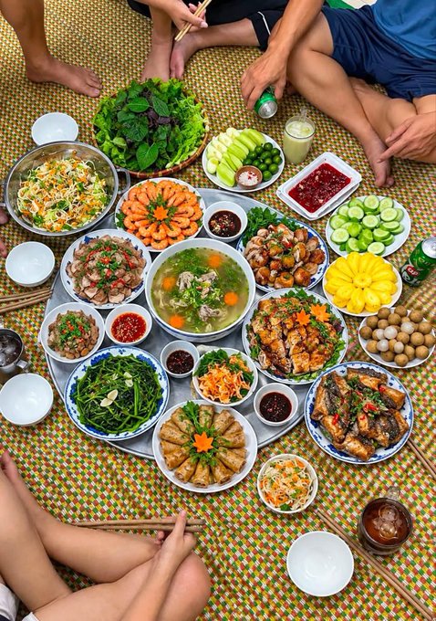 A traditional holiday family feast spread across a woven mat features a circular tray laden with Vietnamese dishes, including spring rolls, grilled meats, soup, fresh herbs, vegetables, and dipping sauces, surrounded by family members sharing the meal together. The caption "CƠM CHIỀU NGÀY LỄ" (Holiday Dinner) reflects the festive gathering of loved ones coming together over an abundant home-cooked meal.