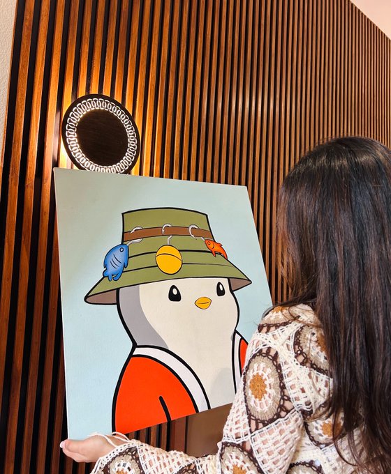 A woman with long dark hair, viewed from behind, holding a cartoon-style painting of a duck character wearing a bucket hat decorated with small fish, set against a wood-paneled wall with a circular decorative mirror.