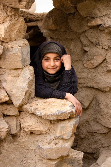 A portrait of a smiling young girl wearing a black headscarf, peering out from a window made of rough, stacked stones.