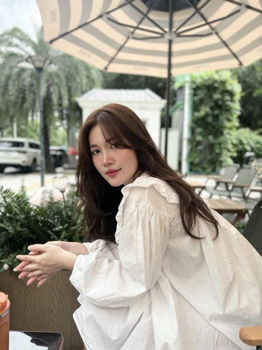 A woman with long brown hair wearing a white blouse, sitting outdoors at a cafe under a striped umbrella with greenery in the background.