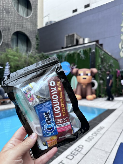 A hand holds a Liquid I.V. Hydration Multiplier electrolyte drink mix pouch by a pool, with a brown bear statue and 'NO DIVING' visible in the background.