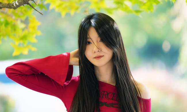 A portrait of a young woman with long black hair wearing a red off-shoulder top against a green leafy background.
