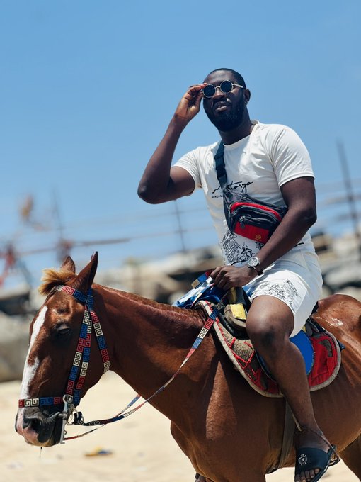 A man wearing sunglasses, a white t-shirt, and a crossbody bag sits on a brown horse on a sunny day, adjusting his sunglasses with his hand.