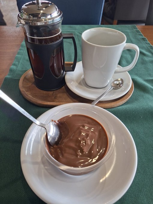 A close-up photo of a French press filled with coffee next to a white mug, and a bowl of chocolate pudding or dessert topped with chocolate syrup on a green placemat.