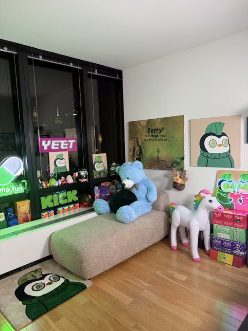 A cozy trading nook by a window overlooking the city features a blue Care Bear seated on a beige bench, surrounded by playful decorations including "YEET" and "KiCK" neon-style signs, a "Sorry! The bridge you water is sold out." art print, and a plush unicorn, perfectly capturing the whimsical "dream setup to trade from."