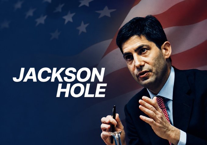 A man in a suit speaks while holding a pen, set against a backdrop of the American flag, with "JACKSON HOLE" displayed prominently in white text, illustrating the central bankers' symposium referenced in the post about its potential impact on BTC's price rally.