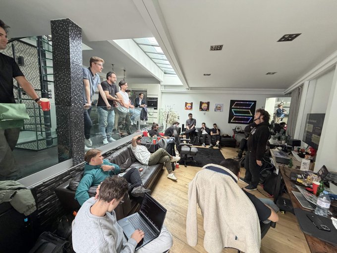 A group of casually dressed young men gathered in a large living room, with some sitting on couches using laptops and others standing, creating a relaxed social atmosphere.
