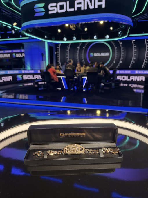 A commemorative 'Breakpoint 2026' champion watch displayed in its presentation box on a reflective black surface, with the Solana-branded conference stage and panel discussion visible in the background.