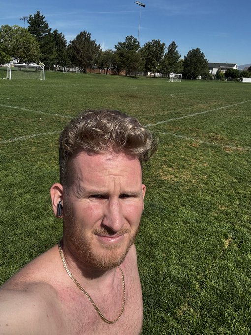 A shirtless man with a beard takes a selfie on a grassy soccer field on a sunny day.