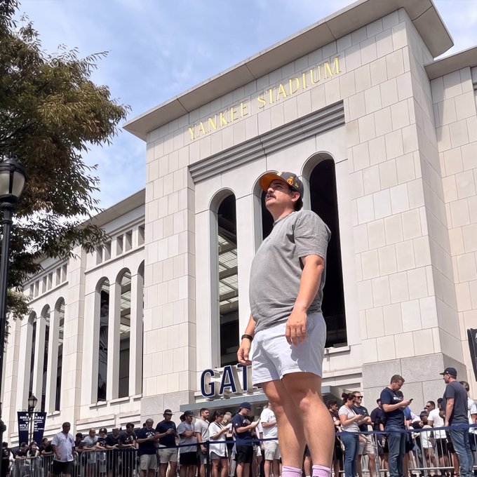 A person wearing an "LA" cap and gray t-shirt standing outside Yankee Stadium, with crowds and stadium signage visible.