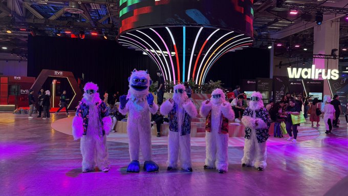 Four people in yeti or abominable snowman mascot costumes stand on a brightly lit convention floor with colorful overhead displays.