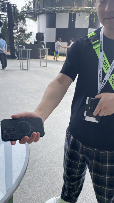 A person wearing a black t-shirt and a lanyard with "IDEXO" and "FN" text holds a black smartphone featuring a circular magnetic ring on its back, standing outdoors at what appears to be an event.