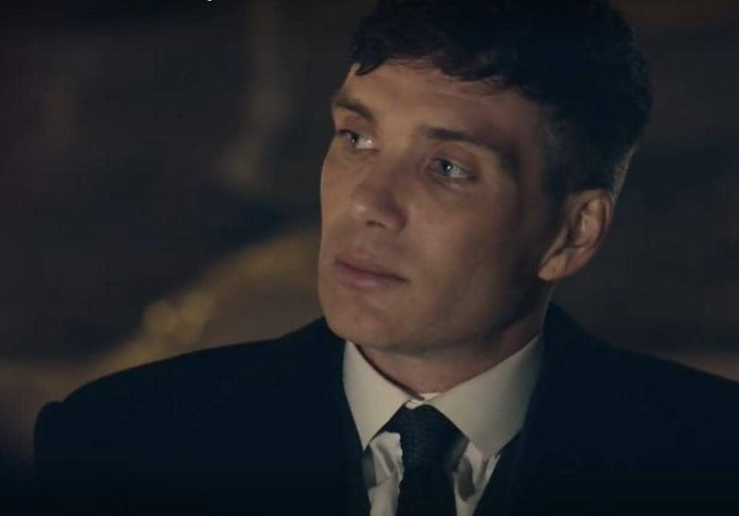 A close-up portrait of a man in a dark suit, white shirt, and dark tie, with short dark hair, looking off-camera with a serious expression in dimly lit surroundings.