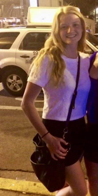 A young blonde woman in a white t-shirt and black skirt smiles at the camera while holding a black purse, standing on a street at night with a taxi in the background.