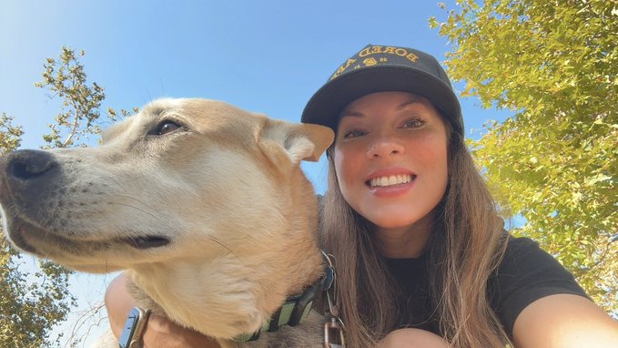 A smiling woman wearing a black 'U.S. AIR FORCE' cap takes a selfie outdoors with a light-colored dog against a backdrop of green trees and blue sky.