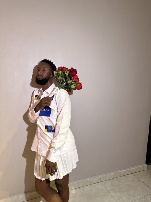 A man poses holding a bouquet of red roses, wearing a white shirt with sponsor logos and a white pleated skirt.