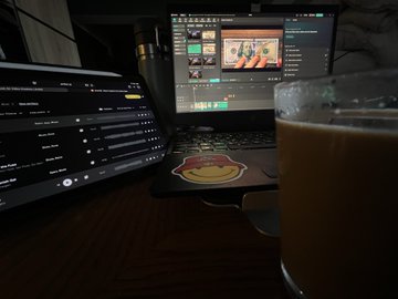 A dimly lit workspace showing a laptop with video editing software open, a tablet with a settings menu on the left, and a glass of coffee in the foreground.