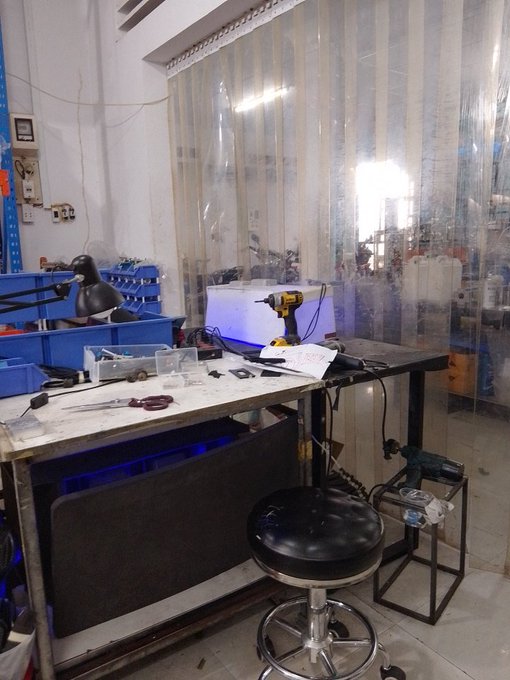 A cluttered workshop bench with a yellow DeWalt drill, scissors, various small tools, and plastic storage bins, with a black padded stool and plastic strip curtains in the background.