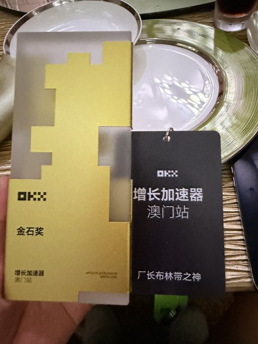A close-up of a gold 'OKX' award plaque reading '金石奖 增长加速器 澳门站' alongside a black 'OKX' card reading '增长加速器 澳门站 厂长布林带之神', placed on a table near gold plates.