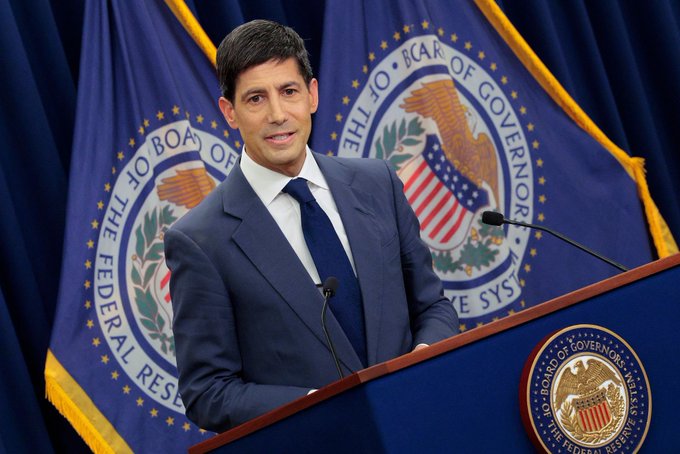A man in a navy suit and tie stands at a podium with the seal of the Board of Governors of the Federal Reserve System, speaking in front of two official flags.