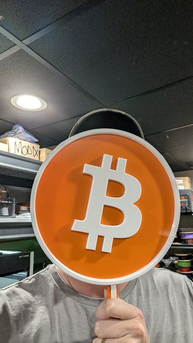 A person holds up a large circular orange sign featuring the white Bitcoin "₿" logo in front of their face, with a "Moody" box visible on a shelf in the background.