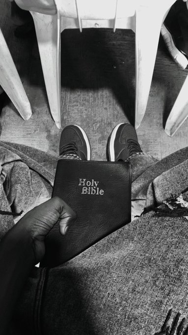 A black-and-white photo shows a person's legs in sneakers as their hand holds a small black book labeled 'Holy Bible' above their lap.