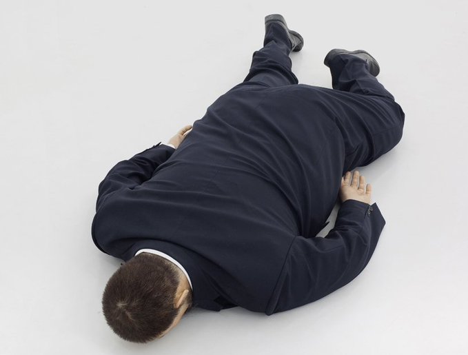 A man in a dark blue suit lies face down on a white floor, with his arms extended outward.