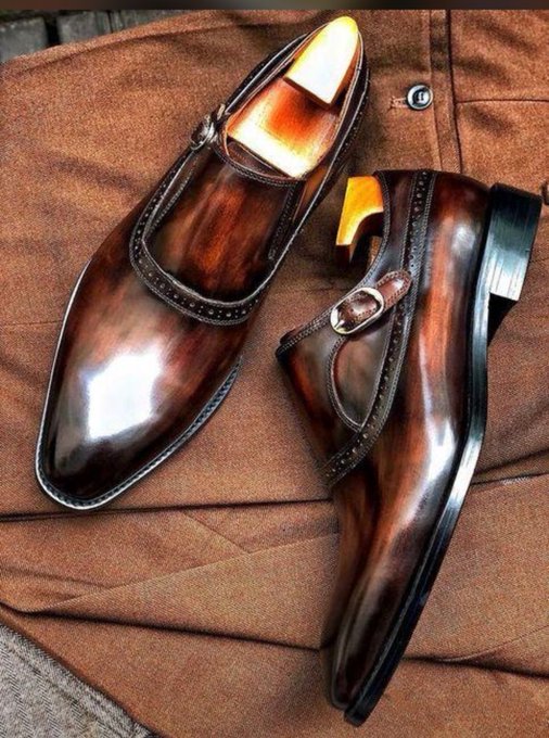 A pair of polished brown leather monk strap dress shoes rests on a brown fabric surface, fitted with wooden shoe trees.