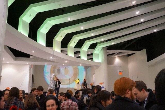 A crowded indoor event space features a stage with a large projection of a soccer ball and Pikachu, beneath a ceiling with distinctive green-lit angular architectural panels.