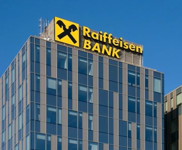 A modern glass office building with a prominent yellow "Raiffeisen BANK" sign and logo on its rooftop against a clear blue sky.