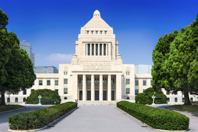 A grand, symmetrical neoclassical building with a tall central tower and columns stands behind a walkway lined with manicured hedges and large green trees under a clear blue sky.