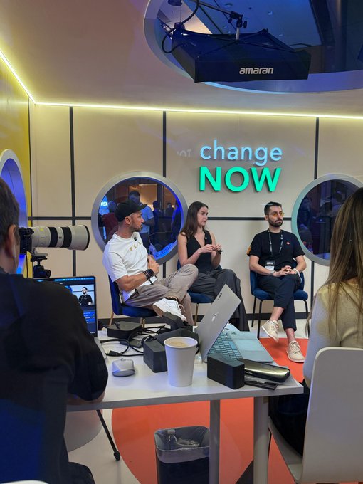 A group of four people sit on stage during a panel discussion at a TOKEN2049 podcast zone, with a camera operator in the foreground and a laptop, microphone, and cup on the table. A large neon sign on the wall reads “change NOW,” and an overhead projector is branded “amazon,” matching the post’s description of live interviews, panels, and conversations from the event floor.