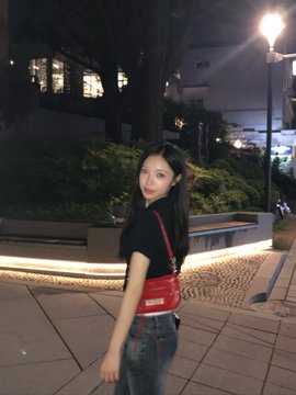 Nighttime photo of a young woman with long dark hair wearing a black top and jeans, carrying a red purse in an outdoor plaza.