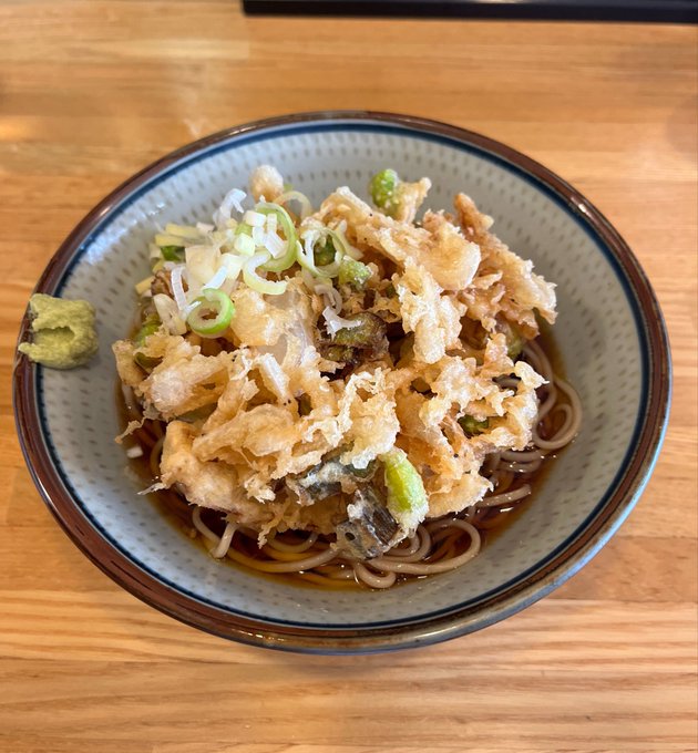 A bowl of Japanese soba noodles topped with a generous portion of kakiage (mixed tempura), chopped scallions, and a side of wasabi in a dashi broth on a wooden table.