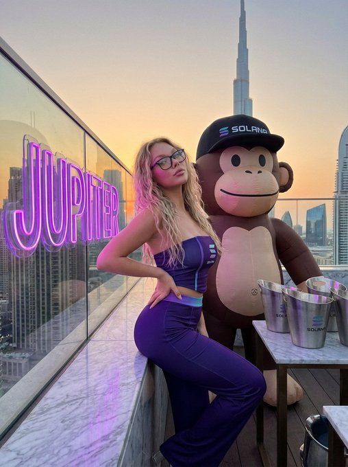 The image features a woman posing with a large inflatable monkey mascot, likely from the company Jumbo. The monkey is positioned behind her, creating an interesting composition for the photo. In addition to the monkey mascot, there are several buckets of beer placed on a table nearby, suggesting that this scene takes place at a social event or gathering.