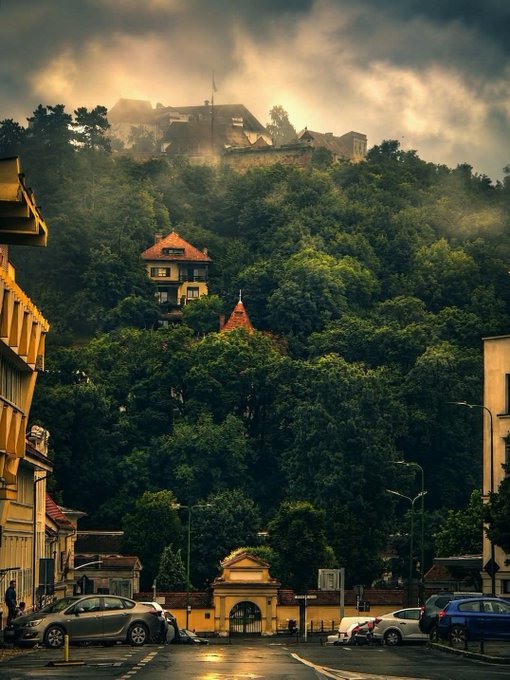 A moody street view looking up a hill toward a fog-shrouded castle or large building, with parked cars and an old town in the foreground.