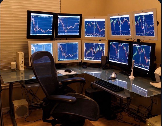 Home office trading setup with six monitors displaying stock charts and candlestick patterns arranged around a glass desk with an ergonomic chair.