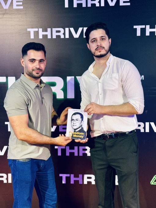 Two men stand together in front of a "THRIVE" branded backdrop, holding up a copy of "Freedom of Money"—a book by Binance founder CZ (Changpeng Zhao), as noted in the caption where the recipient thanks Noman for the gift and expresses excitement to read about CZ's journey from zero to building the world's largest crypto exchange.