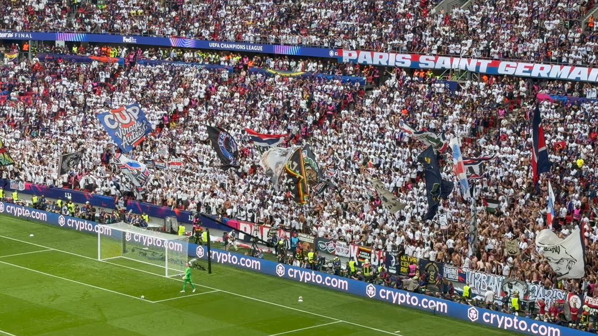 The image shows a large soccer stadium filled with fans, all wearing white clothing. The crowd appears to be watching a soccer match or game taking place on the field. In the foreground of the photo, there are two flags prominently displayed - one red and one blue. These flags may represent different teams or countries participating in the event.
