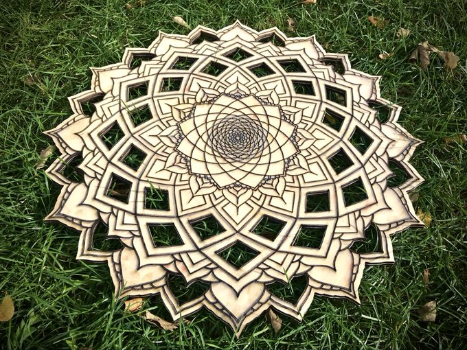 An intricate wooden mandala or lotus-style decorative piece with detailed geometric and floral cutout patterns, resting on green grass.