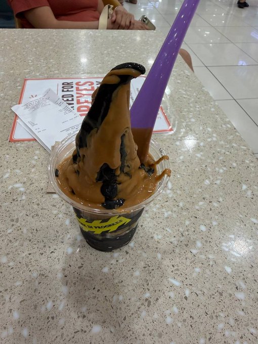 A plastic cup filled with a dessert that looks like a melted popsicle, with a purple stick sticking out, sitting on a speckled countertop near a receipt.