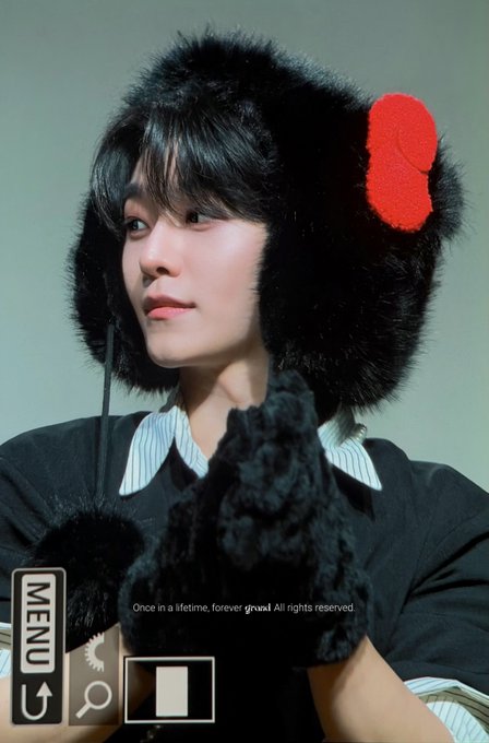A person wearing a fluffy black hat with a red accent and matching black fuzzy gloves poses with hands together, wearing a striped shirt and dark jacket. The image features UI elements with "MENU" text and "Once in a lifetime, forever grow! All rights reserved," suggesting this is a promotional photo for XINLONG (씬롱), likely from their AlphaDrive One album "ALD1."