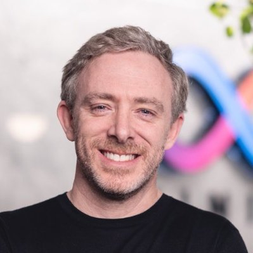 The image shows a smiling man with short graying hair and a beard, wearing a black shirt, who appears to be Dominic Williams, the founder of Dfinity and creator of the Internet Computer Protocol. The post references him in connection with the protocol successfully defending against a recent wave of attacks.