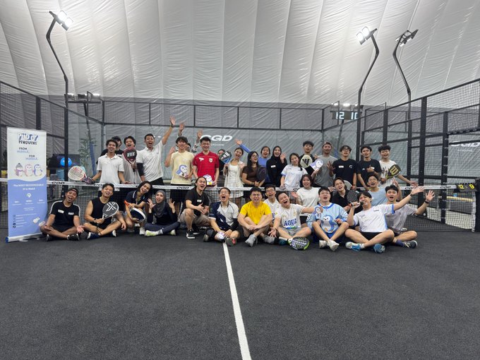 A group photo of approximately 25 people posing together on an indoor padel court under a domed structure, holding padel rackets and cheering.