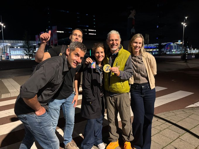 Group photo of five people smiling and posing together at night on a city street, with two holding up small items.