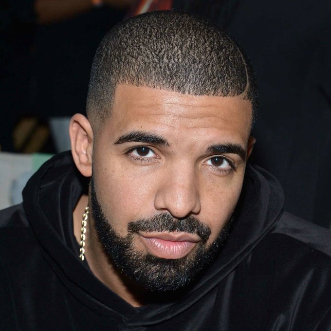 A close-up portrait of a man with short hair, a beard, and a gold chain wearing a black hoodie.