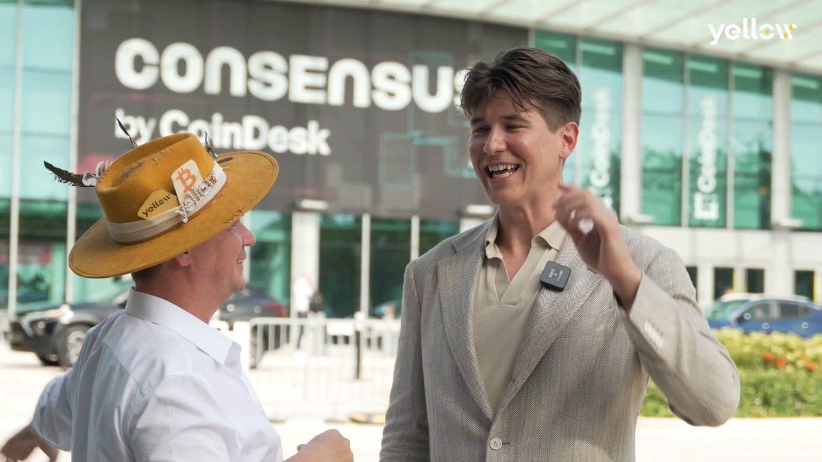 The image features two men standing outside, one wearing a yellow hat and the other dressed in a suit. They are engaged in conversation with each other, possibly discussing something related to cryptocurrency or financial technology. The background of the photo shows a building, which could be an office or a store associated with the company CoinDesk.