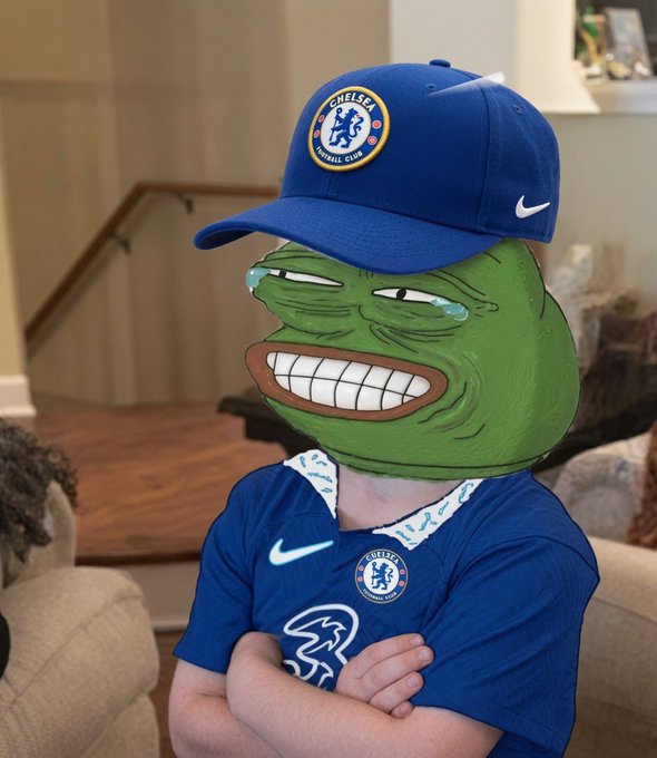 A person wearing a blue Chelsea FC jersey and cap with a green Pepe the Frog face superimposed over their head, standing with arms crossed in a living room setting.