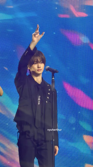 A young man on stage in a black jacket raises his arm toward a microphone stand against a blue screen, with 'yyuharthur' visible on the image.