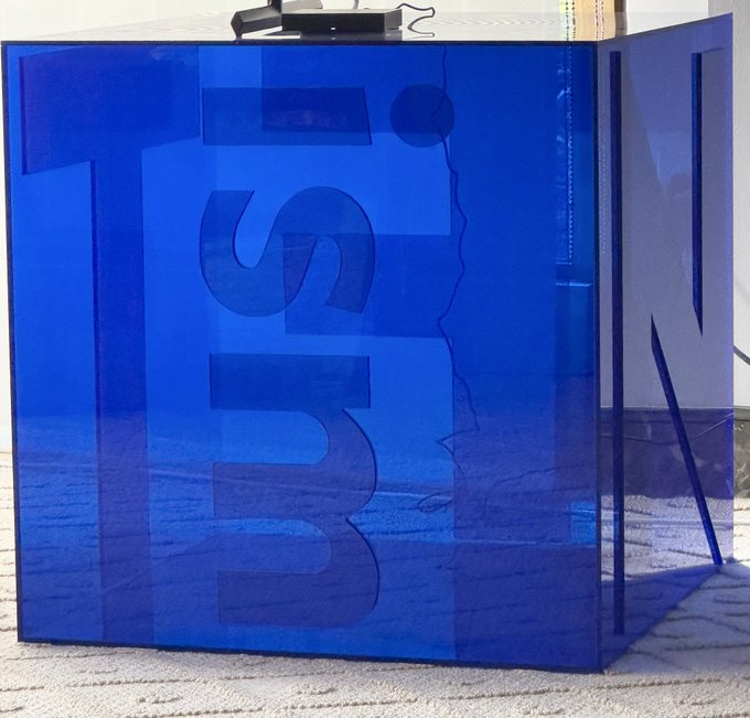 A translucent blue acrylic or glass box with the word 'TURN' embossed on its sides sits on a textured carpet.